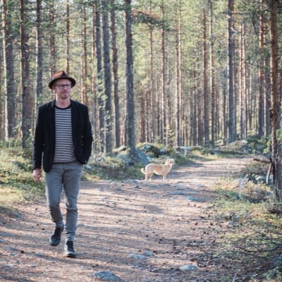 Mies hattu päässä, pikkutakki päällä ja farkut jalassa metsäisellä polulla, taustalla kellertävä koira