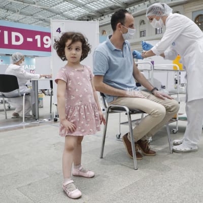 En sittande man får coronavaccin på en klinik, hans lilla dotter står bredvid.