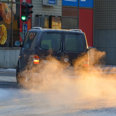 Bil kör på snötäckt väg och lämnar ett moln av avgaser bakom sig. 