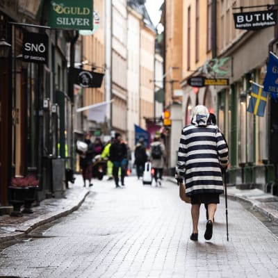 Kvinna i randig kappa går i gamlastan i Stockholm. Gatan är folktom och kvinnan går med ryggen mot kameran.