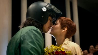 Mies musta moottoripyöräkypärä päässa ja punatukkainen nainen kukkakimppu kädessä ovat puolilähikuvassa juuri suutelemaisillaan, kuva elokuvasta Annette.