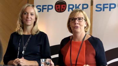 Två kvinnor står vid höga bord. I bakgrunden en nyckelpiga och  texten SFP RKP. Från vänster, riksdagsledamoten Sandra Bergqvist (SFP) och Anna-Maja Henriksson som är är Svenska Folkpartiets ordförande.