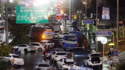 Bilar och bussar står huller om buller i vatten längs en större gata medan regnet fortsätter att ösa ner. Det är mörkt och ljuset från vägbelysning speglar sig i vattnet. På skyltarna står destinationer både på koreanska och med västerländska bokstäver.