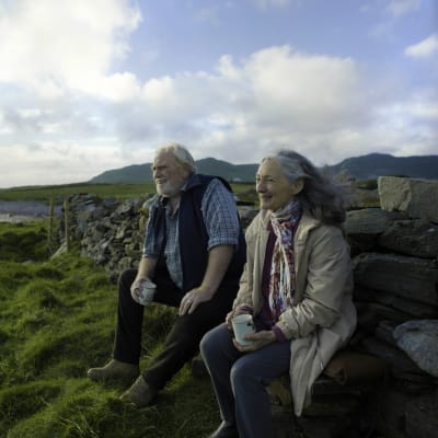 En leende Howard (James Cosmo) och Annie (Bríd Brennan) sitter vid en stenmur och ser ut över havet, bakom dem syns gröna vidder och berg.