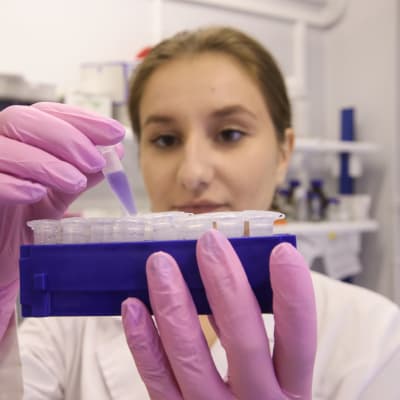 Kvinna i vid laboratorierock och gummihandskar håller i ett blått kärl med små plastbehållare. Det ryska coronavaccinet tillverkas vid det ryska nationella institutet för epidemiologi och mikrobiologi samt vid läkemedelsfabriker i Ryssland.