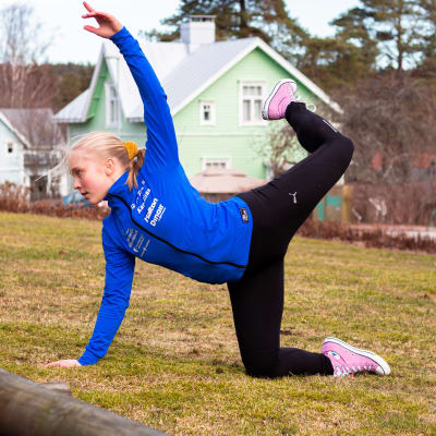 En ung kvinna med blå sportjacka gör en gymnastisk rörelse på en gräsmatta.Ett ben och ena armen i luften. de andra mot marken. Hus i bakgrunden