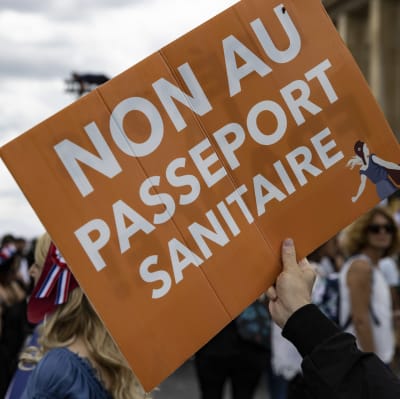 Bild på en orange skylt där det står "nej till hälsopass" skrivet på franska. Bakom syns en folkmassa och Eiffeltornet.