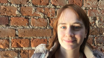 Ailana Moore, en ung kvinna med brunrött halvlångt hår och en beige jacka stående framför en tegelvägg, studerar folkloristik vid Åbo Akademi.