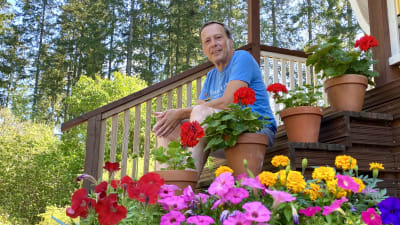En medelålders man i shorts och t-skjorta sitter på en trappa. Bilden är tagen från sidan och nerifrån så att blommorna på trappan syns i förgrunden.