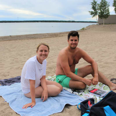 En kvinna och en man sitter på var sin handduk på en sandstrand.