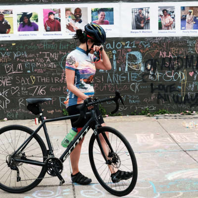 Pyöräilijä peittää silmänsä kädellään. Hänen takanaan on seinä, jossa on rivissä kymmenen ihmisen kuvat ja nimet.