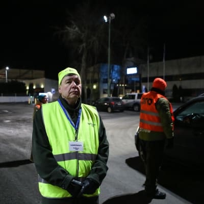 Lakkovahtina toimiva mies seisoo pimeässä keltainen liivi päällään. Takana on kaksi muuta miestä.