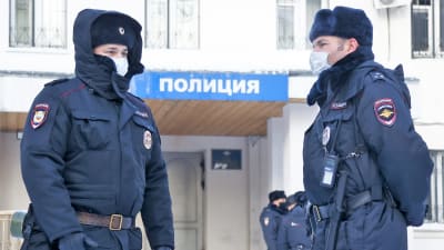 Kaksi poliismiestä seisoo poliisiaseman edustalla. Molemmilla on karvalakit päässä, toinen on nostanut hupun lakin päälle. Miehillä on suojamaskit.
