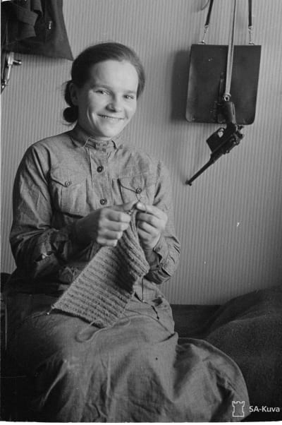 Svarvit bild på en kvinna i lottauniform som sitter på en stol och stickar. På väggen bakom henne hänger ett skjutvapen från en krok.