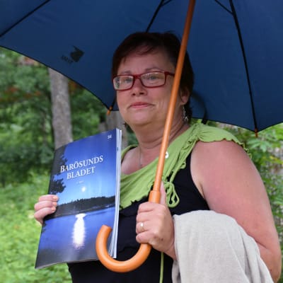 Margita Björklöf står och håller i ett paraply och ett exemplar av Barösundsbladet.