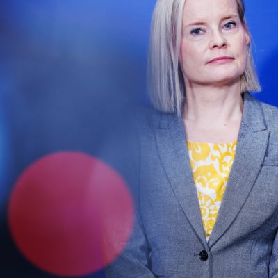 Riikka Purra tittar åt sidan. Hon är klädd i en grå kavaj och gul skjorta under. Hon står vid en blå vägg.