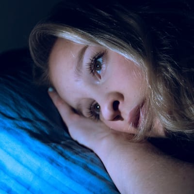 Nuori nainen pää tyynyllä selaa kännykkää unettomana.