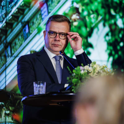 Petteri Orpo rättar till sina glasögon när han står vid en talarstol.