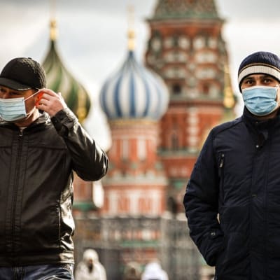 Två män står på Röda torget i Moskva med Vasilijkatedralens lökuloler i bakgrunden. Den ena mannen bär munskydd, den andra tar på sig munskydd.