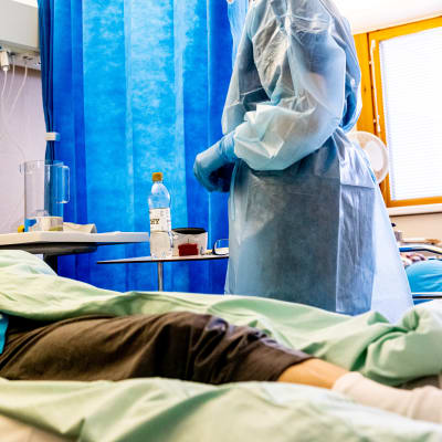 Suojavarusteisiin pukeutunut lähihoitaja Janina Kinnunen keskustelee potilaan kanssa erityshuoneessa.