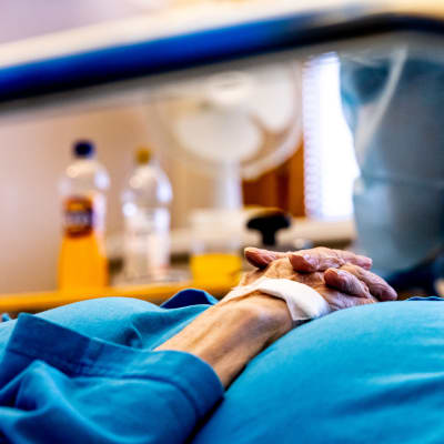 Koronapotilas kädet ristissä erityshuoneessa, suojavarusteisiin pukeutunut hoitaja vieressä.