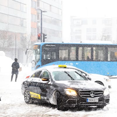Jalankulkijoita, taksi ja bussi lumisateessa.