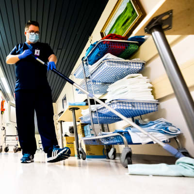 En städare moppar golvet i en sjukhuskorridor. Till höger en rullvagn med handdukar, till vänster en gul sopkorg.