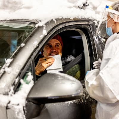 En man sitter i en bil, i färd att snyta sig inför ett coronatest, medan en person i skyddsmundering står bredvid bilen med en testningspinne i handen.