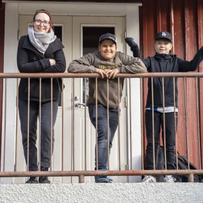 En kvinna och två pojkar lutar mot ett järnräcke på en trappa. Bakom ett rött trähus.