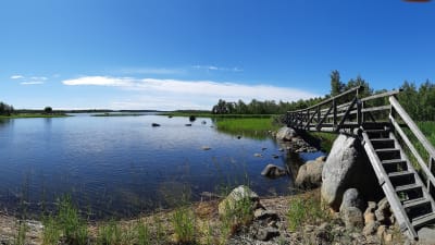 Vatten i skärgård. Det är sommar och himlen är blå. En smal träbro går över en del av vattnet.