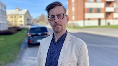 En man i beige kavaj och blå skjorta och glasögon står ute i solen.