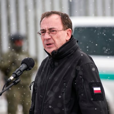 Puolilähikuvassa mustaan takkiin pukeutunut mies puhuu mikrofoniin. Hihassa Puolan lippu. Taustalla maastokuvioiseen uniformuun pukeutunut henkilö.
