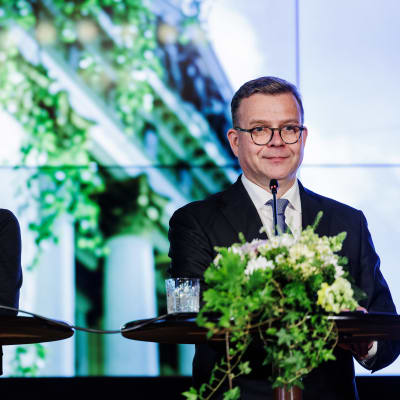 Riikka Purra och Petteri Orpo står vid varsitt blomsterprytt podium med mikrofoner framför sig.