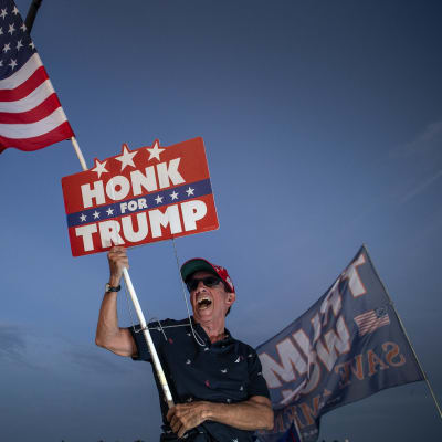 Lippalakkiin ja aurinkolaseihin sonnustautunut mies heiluttaa suurta Yhdysvaltain lippua ja kylttiä tekstillä "Honk for Trump".