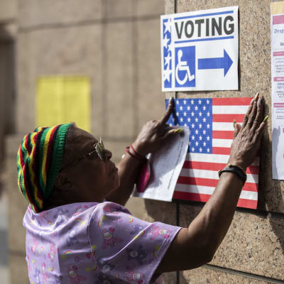 En kvinna hänger upp en poster som föreställer USA:s flagga på en vägg. Ovanom den postern finns en annan poster med texten "rösta".