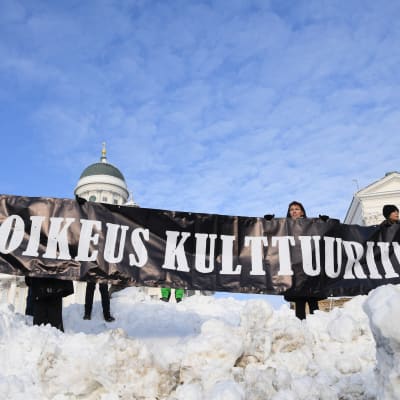 Demonstranter håller upp en banderoll där det står "oikeus kulttuuriin" framför domkyrkan.