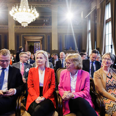 Petteri Orpo, Riikka Purra, Anna-Maja Henriksson och Sari Essayah sitter bredvid varandra i en stor sal, i bakgrunden fler människor.