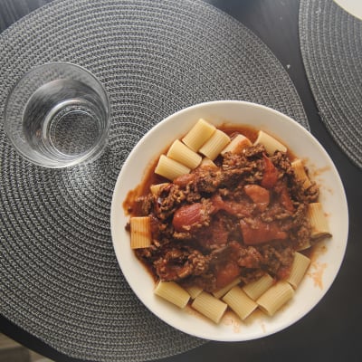 En skål med pasta och köttfärssås står på ett bord med ett glas vatten.