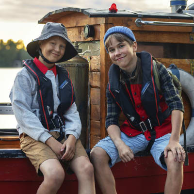 Kaksi poikaa istuu veneen laidalla, katsoo kameraan ja hymyilee.