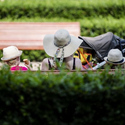 Tre personer sitter på en bänk. De har hattar på huvudet och äter glass i hettan.