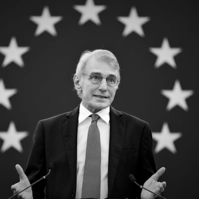 En svartvit bild av David Sassoli som pratar. Han står framför en vägg som har EU:s flagga på sig.