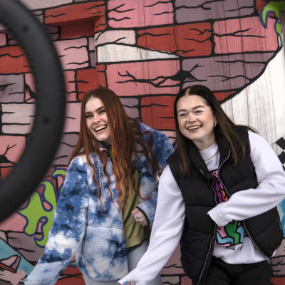 Kaksi nuorta naista nojaavat graffitiseinää vasten. Etualalla on ring light -kuvaustarvike.