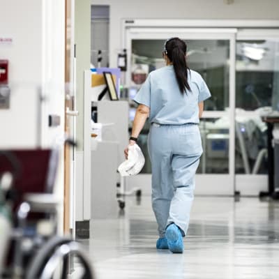 En sjukskötare går i en sjukhuskorridor. Hon har ryggen mot kameran.