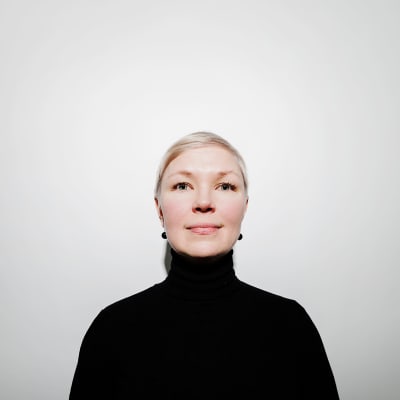 Marjo Rantala står vid en vit vägg. Hon tittar in i kameran. Hon är klädd i en svart polotröja.