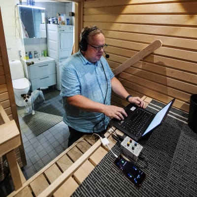 En person står i en bastu vid sin dator med headset på sig, bakom honom en hund.