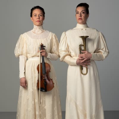 Riikka Ahokas ja Tuuli Saksala poseeraavat valkeissa mekoissa vierekkäin. Ahokkaalla on käsissään viulu ja Saksalalla trumpetti.