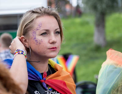 Ung kvinna med smink och glitter i ansiktet, drar sitt hår bakom örat. Hon har en regnbågsflagga draperad över axlarna. Tittar beslutsamt på något utanför bild. 