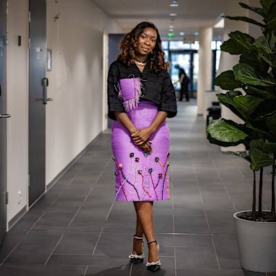 Vanessa Pecani seisoo käytävällä ja poseeraa kameralle. Hänellä on yllään musta paita ja vaaleanpunainen hame, johon on kuvioitu kukkia.