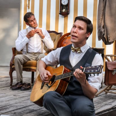 Spelamannen spelar gitarr i förgunden. Bakom honom sitter Agaton och knäpper översta knappen på sin skjorta. De är klädda i gråvita kostymkläder.