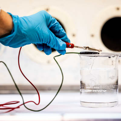 En hand i blå plasthandske håller en liten kabel i ett vattenglas för att genomföra elektrolys.